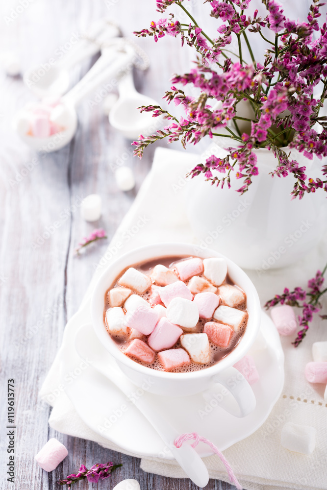 Hot chocolate with mini marshmallows with pink flowers in jug on white wooden background. Hot chocolate with mini marshmallows with pink flowers in jug on white wooden background.