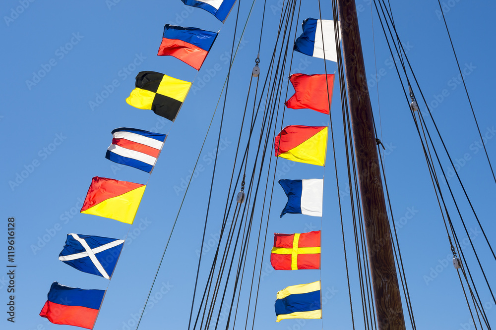 Fototapeta premium Colorful nautical sailing flags flying in the wind from the lines of a sailboat mast backlit in bright blue sky by the sun