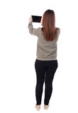back view of standing young beautiful woman using a mobile phone. girl watching. A girl in a gray sweater holds a tablet on outstretched arms.