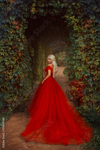 Beautiful blonde girl with long hair. She is in a luxurious red dress. Queen walks near the castle. Vampire passion, gothic story. Smoky eyes and red lips.  