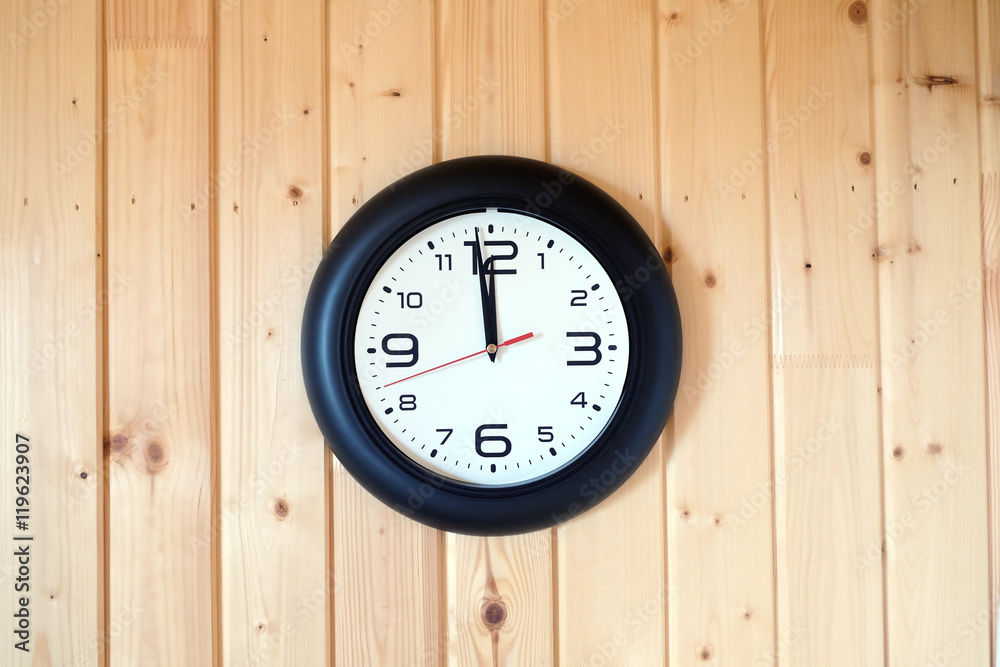 Big round wall clock with a black rim with arrows showing twelve o'clock hangs on brown wooden wall from vertical planks horizontal view indoor on wooden background Big round wall clock with a black rim with arrows showing twelve o'clock hangs on brown wooden wall from vertical planks horizontal view indoor on wooden background