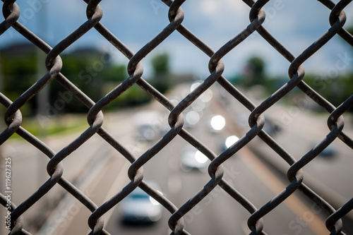 Image of chain link fence on bridge over pass. Chain fence on bridge above highway. Abstract industrial image of chain link fence and cars on highway.