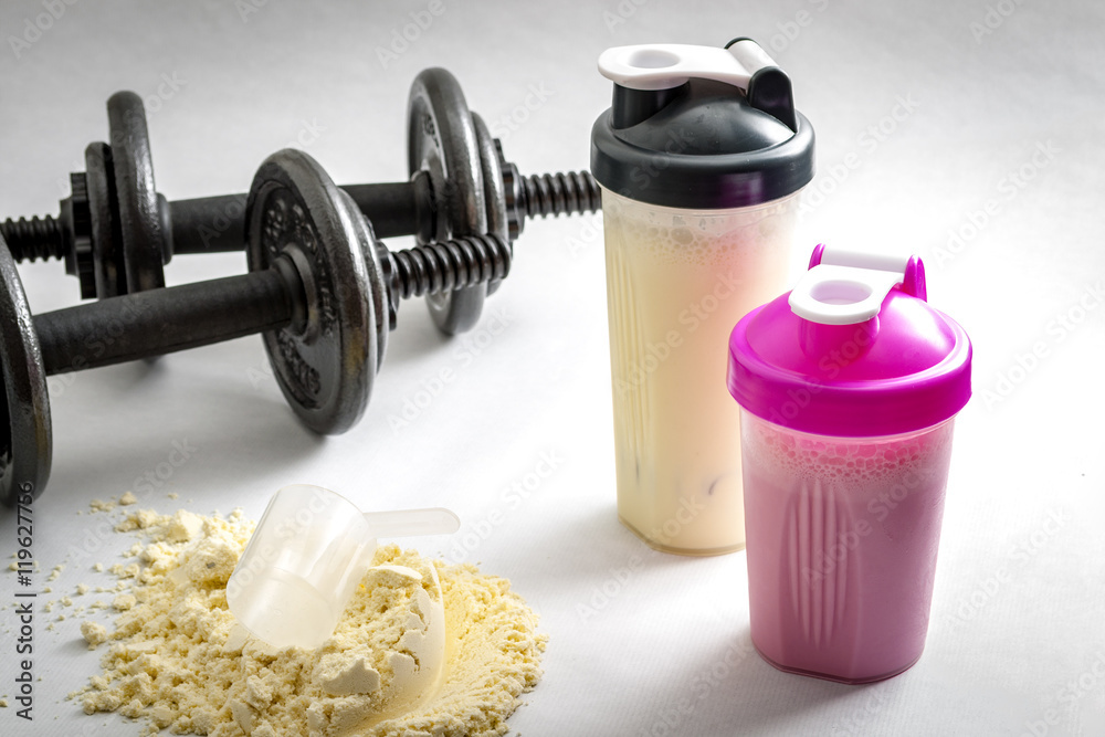 Fitness and workout concept with dumbbells, protein shakers and a scoop in protein powder. The two shakers have black and pink lids and the background is white