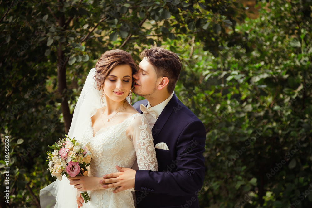 Handsome groom in blue suit hugs dreamy bride while posing in th Handsome groom in blue suit hugs dreamy bride while posing in th