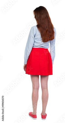 back view of standing young beautiful woman. Long-haired brunette in red skirt looks down sideways.