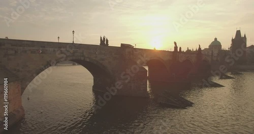 Aerial camera flies fast along the Charles Bridge in Prague as the sun rises behind it. 