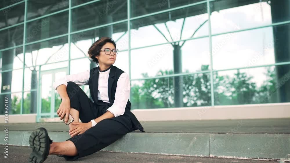 Young beautiful businesswoman sitting on street, business centre background. Slow motion. Young beautiful businesswoman sitting on street, business centre background. Slow motion.