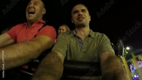 Slow motion of excited scared men enjoying crazy roller coaster ride in the amusement park at night.