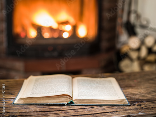 Opened book on the wooden table. Fireplace with warm fire on the
