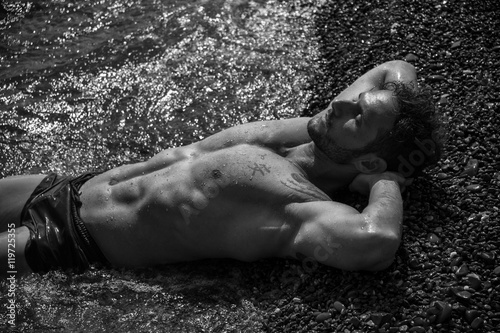 Handsome muscular shirtless man on the beach lying on gravel, with eyes closed