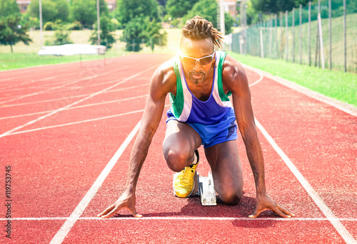 Afro american athlete ready to sprint at starting line - Runner kneeling on blocks of red athletics track - Concept of human concentration at sports competition 