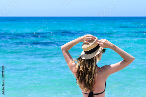 Portrait of young adorable caucasian woman in sunglasses and summer straw hat staying over background of turquoise sea at tropical exotic sandy beach in Sardegna island, Italy