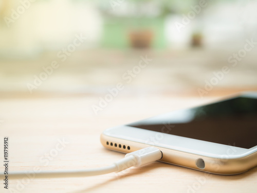 Close-up of white smartphone charging battery with cable on wooden table with copy space and blurry background. Soft light