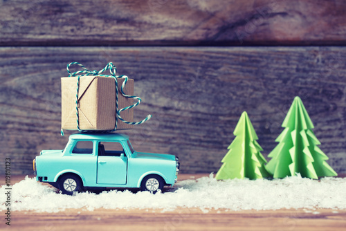Toy car carrying a Christmas gift with snow and paper fir trees, vintage filter