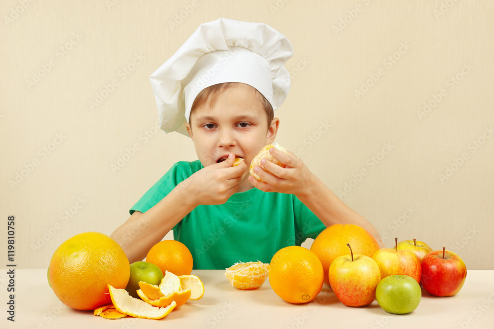 Little boy in chefs hat eat fresh orange at the table with fruits Little boy in chefs hat eat fresh orange at the table with fruits