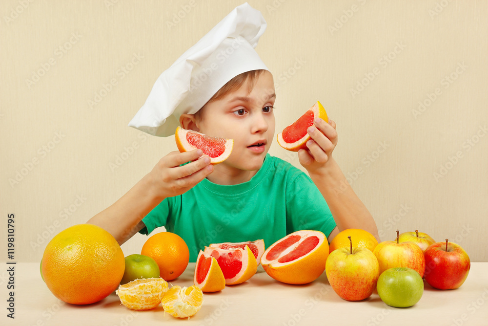 Little kid in chefs hat with two slices of grapefruit at the table with fruits Little kid in chefs hat with two slices of grapefruit at the table with fruits