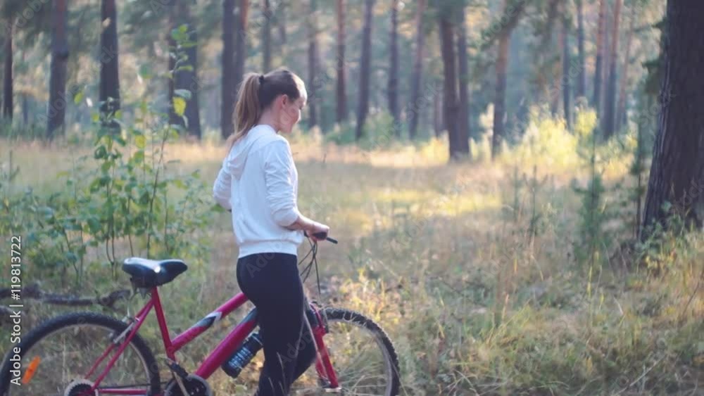 Slow motion. Young athletic woman walking in the forest with bicycle. Sporty woman in white jacket in forest. Slow motion. Young athletic woman walking in the forest with bicycle. Sporty woman in white jacket in forest.