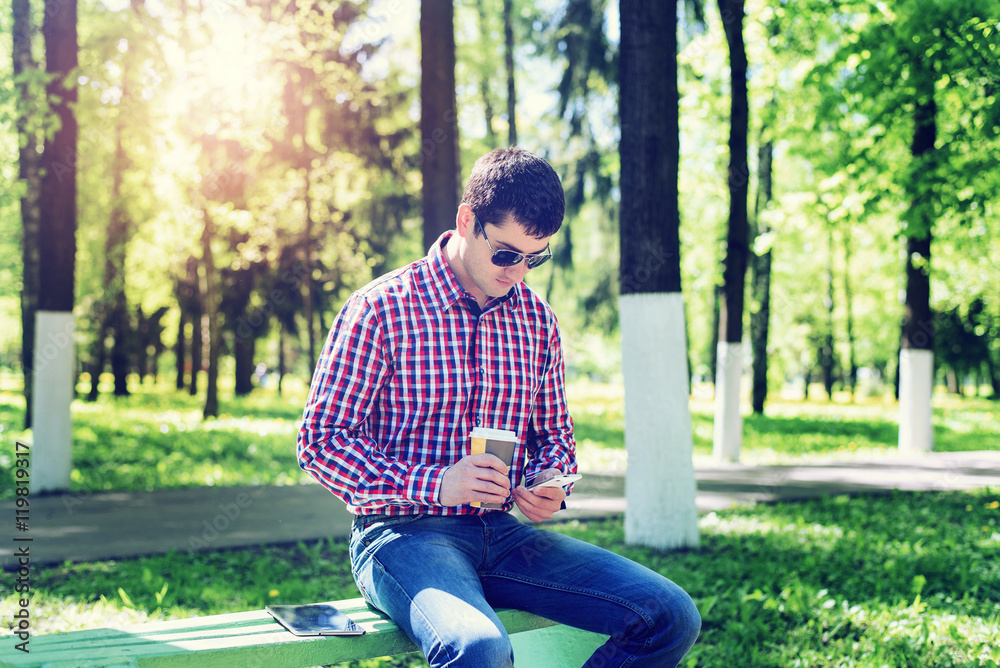 The man in the summer in the park, relaxing with a cup of coffee or tea, reading sms on phone wearing glasses, news feeds in social networks, in jeans bright sunny day. Idea concept businessman The man in the summer in the park, relaxing with a cup of coffee or tea, reading sms on phone wearing glasses, news feeds in social networks, in jeans bright sunny day. Idea concept businessman
