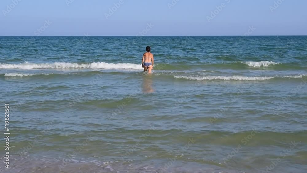 Young slim woman in swimsuit and short black hair goes into water of sea or ocean slender Young slim woman in swimsuit and short black hair goes into water of sea or ocean slender