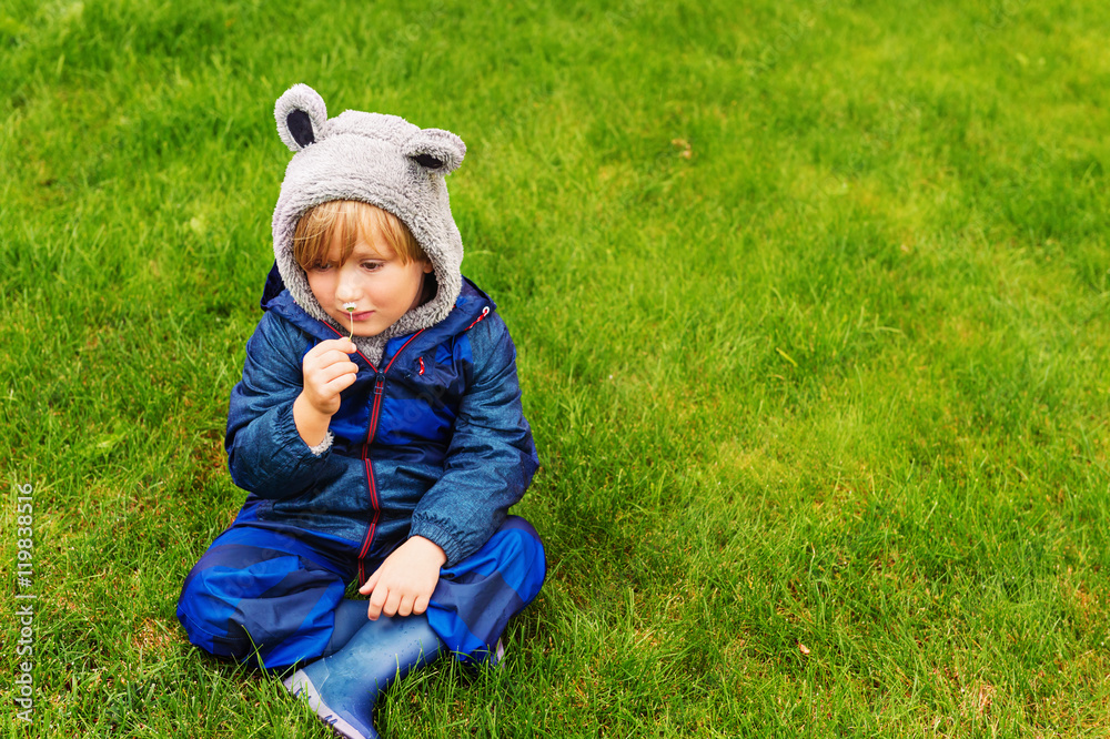 Cute kid resting outdoors, sitting on a lawn, playing on a rainy day, wearing blue waterproof all-in-one suit Cute kid resting outdoors, sitting on a lawn, playing on a rainy day, wearing blue waterproof all-in-one suit