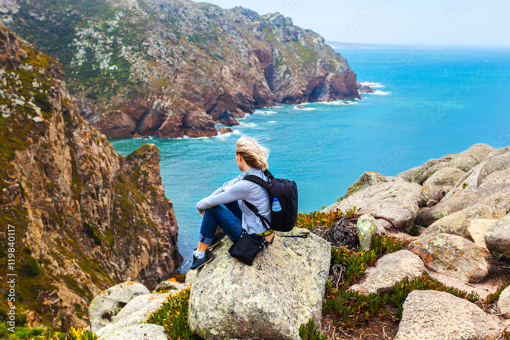 Female traveler sitting on Cape Roca against the background of the Atlantic Ocean, Sintra, Portugal