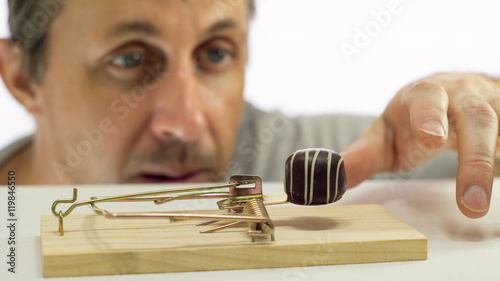 Close up shot of a man looking eagerly at eating a sweet chocolate that is in a mouse or rat trap which indicates that he should not eat it or is on a diet. photo