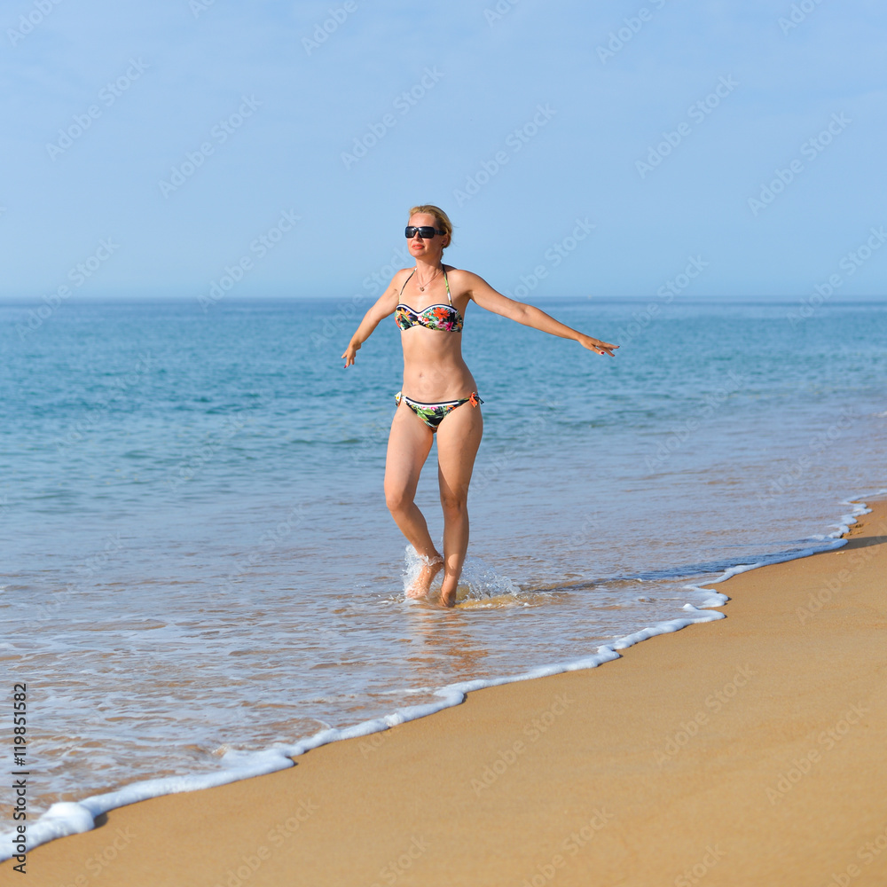 Portrait of attractive woman in bikini and sunglasses holding arms wide happy and joyful on beach holiday, sunny blue sky outdoors background