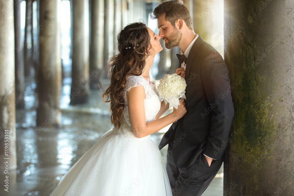 Naklejka premium Beautiful newlywed couple intimate passionate kiss golden hour sunset pier beach