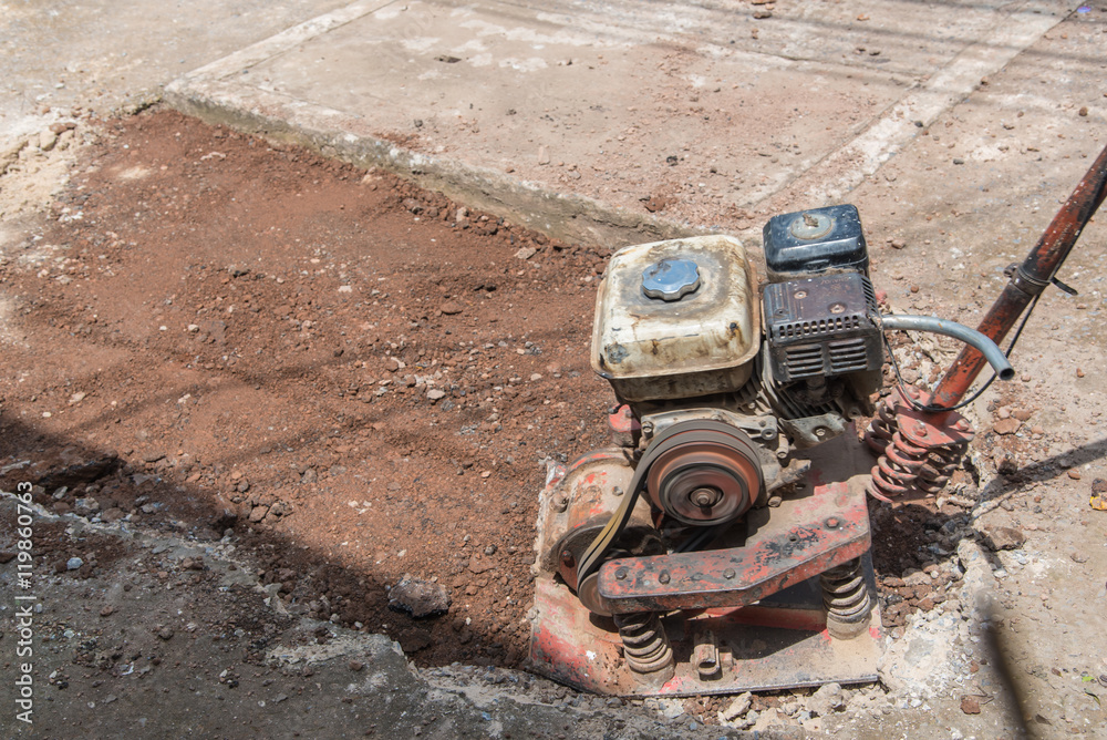 Worker reversible plate compactor on a roadwork site. Excavator in background.Repair cracked Sidewalk Worker reversible plate compactor on a roadwork site. Excavator in background.Repair cracked Sidewalk