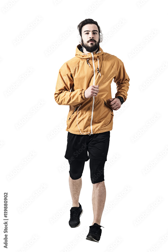 Determined young jogger in jacket with headphones running towards camera. Toned desaturated full body length portrait isolated on white studio background. Determined young jogger in jacket with headphones running towards camera. Toned desaturated full body length portrait isolated on white studio background.