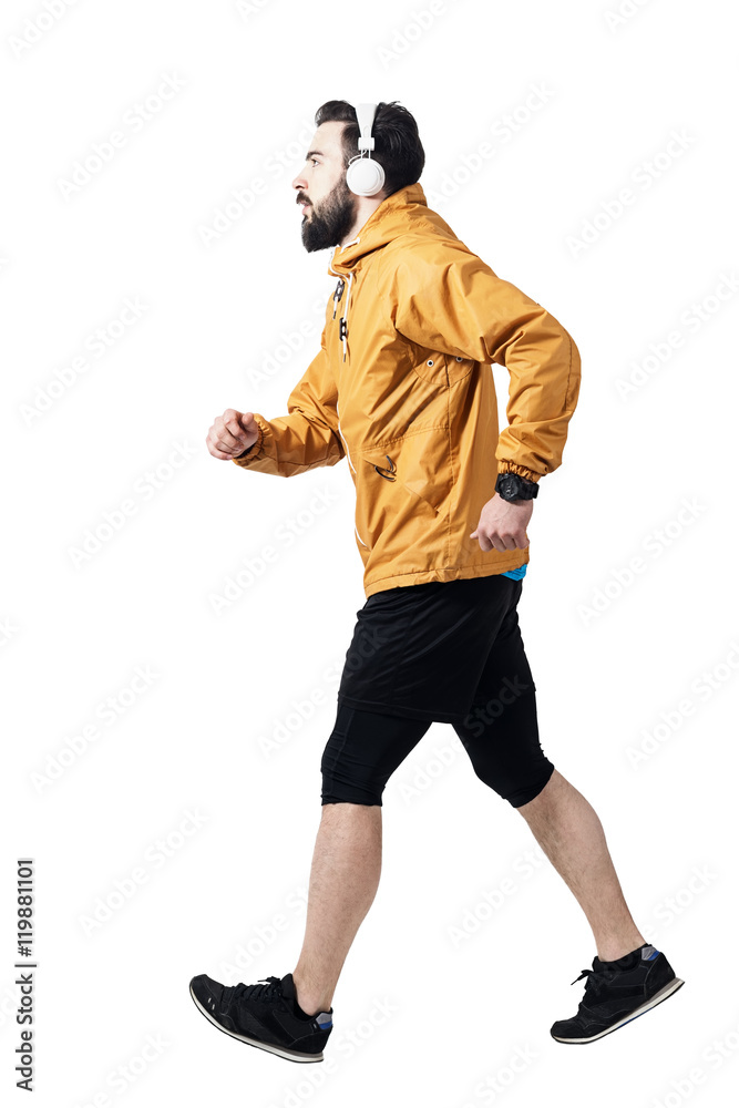 Side view of athlete jogging with headphones wearing jacket. Toned desaturated full body length portrait isolated on white studio background. Side view of athlete jogging with headphones wearing jacket. Toned desaturated full body length portrait isolated on white studio background.