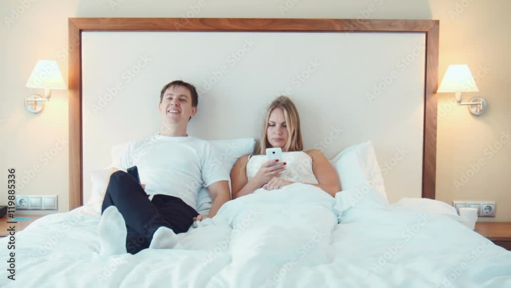Laughing man in blue jeans making himself comfortable in bed with woman using phone at hotel room Laughing man in blue jeans making himself comfortable in bed with woman using phone at hotel room
