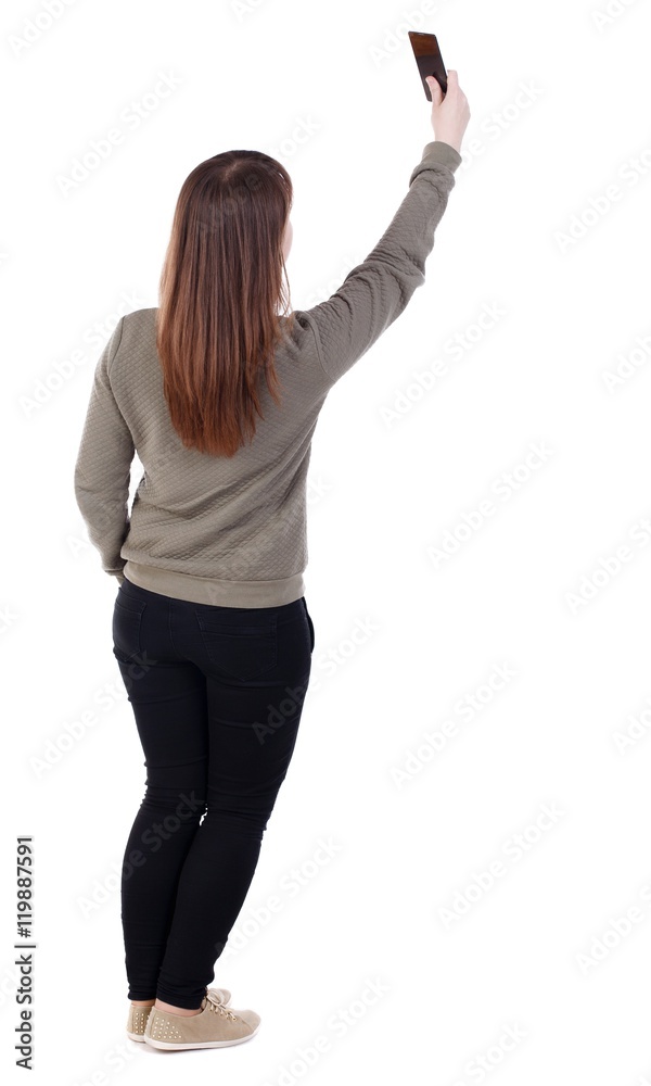 back view of standing young beautiful woman using a mobile phone. girl watching. girl is standing back and top phone photographs themselves. back view of standing young beautiful woman using a mobile phone. girl watching. girl is standing back and top phone photographs themselves.