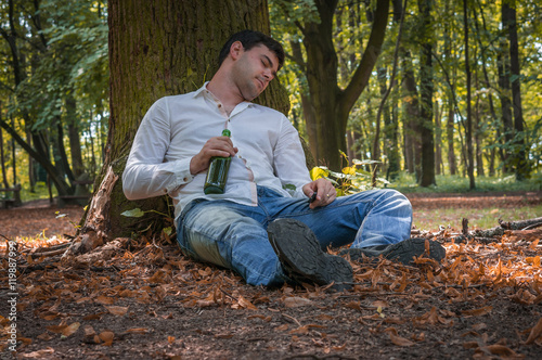 Drunk man lies on the ground under the tree with a beer bottle