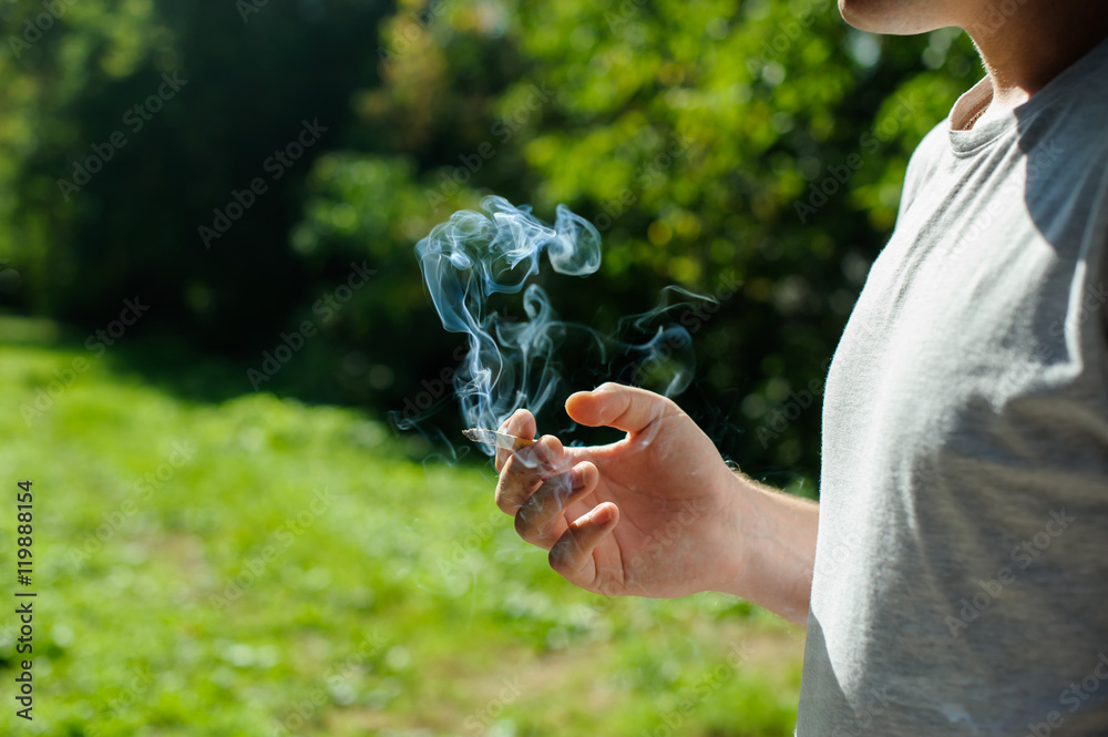 a young man dressed in light and dark ftbolku cap, smoking a cigarette emitting smoke in the background of summer green sunny day