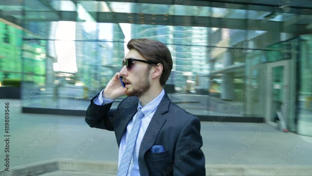 Handsome businessman in sunglasses is talking on the phone near the modern business center, skyscraper. The camera is moving around man (steadicam shot) Handsome businessman in sunglasses is talking on the phone near the modern business center, skyscraper. The camera is moving around man (steadicam shot)