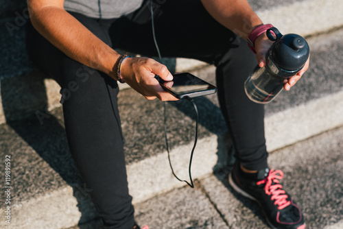 Sport fitness female sitting on stone stairs resting and listening music on phone with bottle of water while resting from outdoors urban training exercise workout. Woman wellness concept.