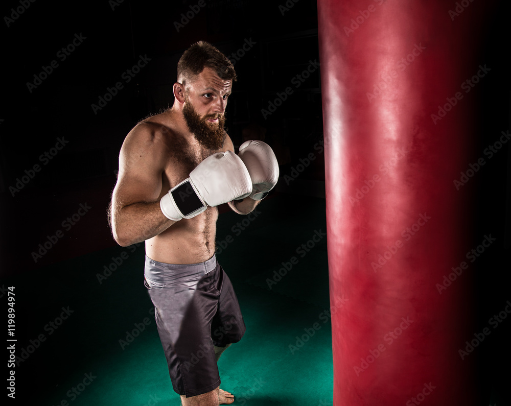 Muscular hipster fighter giving a forceful kick during a practise with a boxing bag.