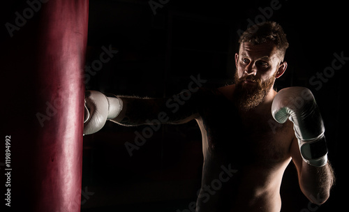 Muscular hipster fighter giving a forceful kick during a practise with a boxing bag. © Lukas Gojda