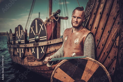 Viking warrior with sword and shield standing near Drakkar on the seashore. photo