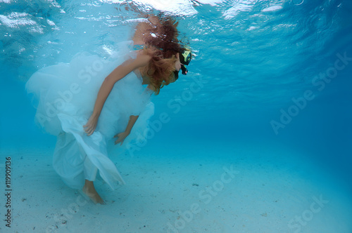 Young beautiful woman with long hair in a long white wedding dress in mask looking under water, underwater wedding in the Indian ocean, Maldives