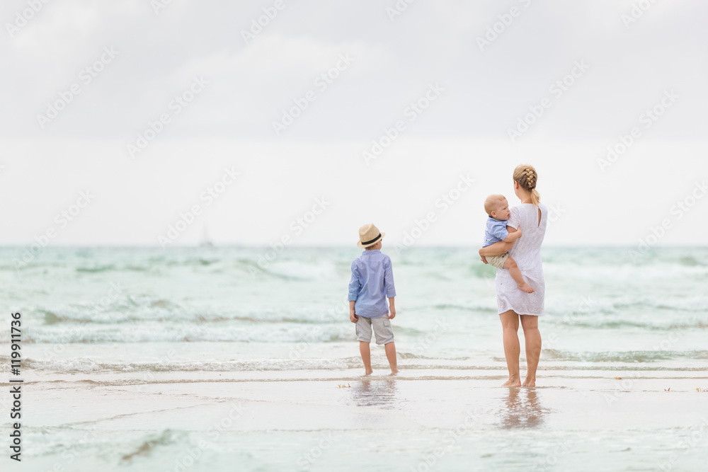 Young mother in white dress walking with her two little boys along the ocean beach. Woman with baby and boy enjoying vacation by the sea. On the empty beach. Motherhood. Water background. copy space.