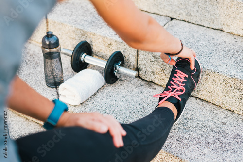 Sport athlete runner female stretching leg on urban stone stairs during training exercise workout routine with bottle of water, towel and dumbbell. Fitness woman outdoor routine.