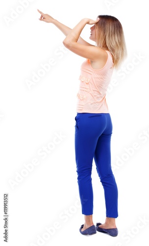 Back view of pointing woman. Isolated over white background. Blonde in blue pants looks into the distance and points a finger.