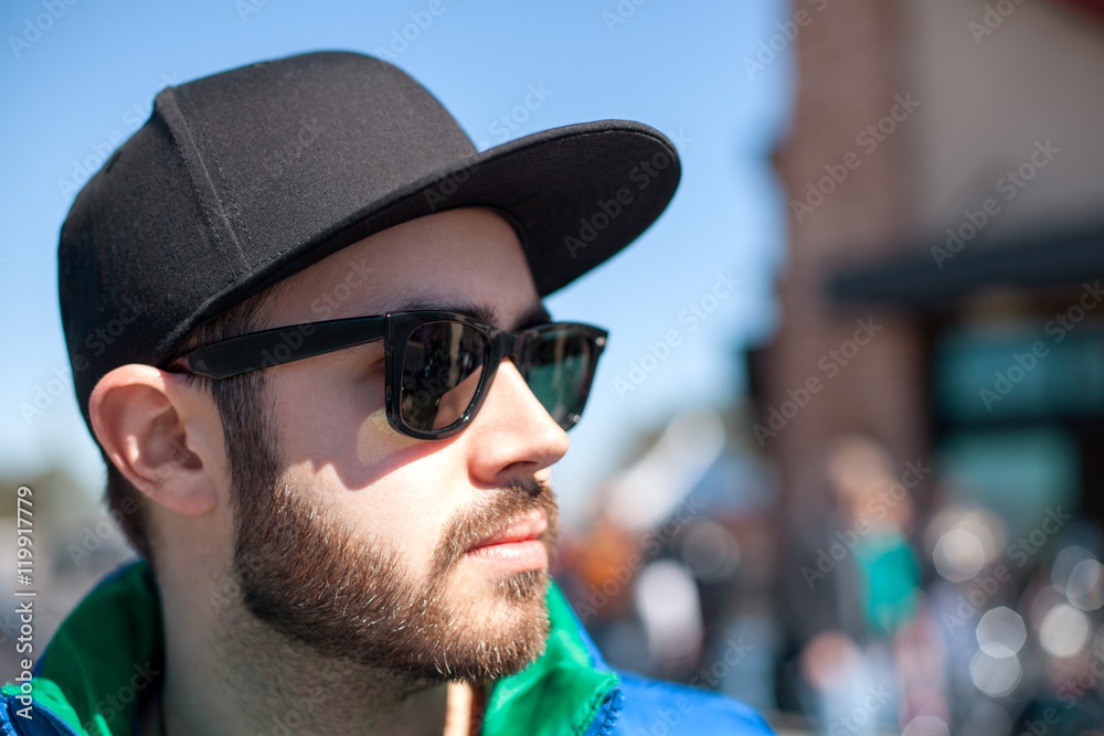 Fototapeta premium Portrait of young handsome man with black beard wearing dark sunglasses, cap and blue sleeveless jacket looking at the street on outdoor in the summer day