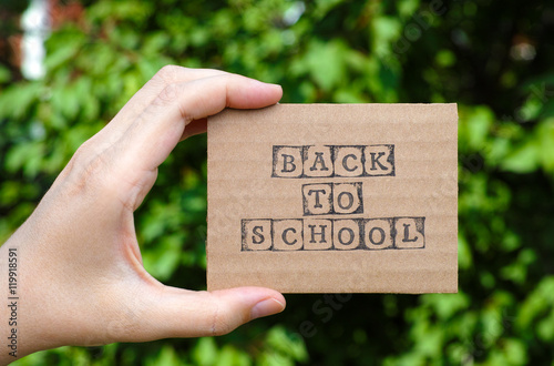 Woman hand holding cardboard card with words Back to School made by black alphabet stamps opposite green floral background. 