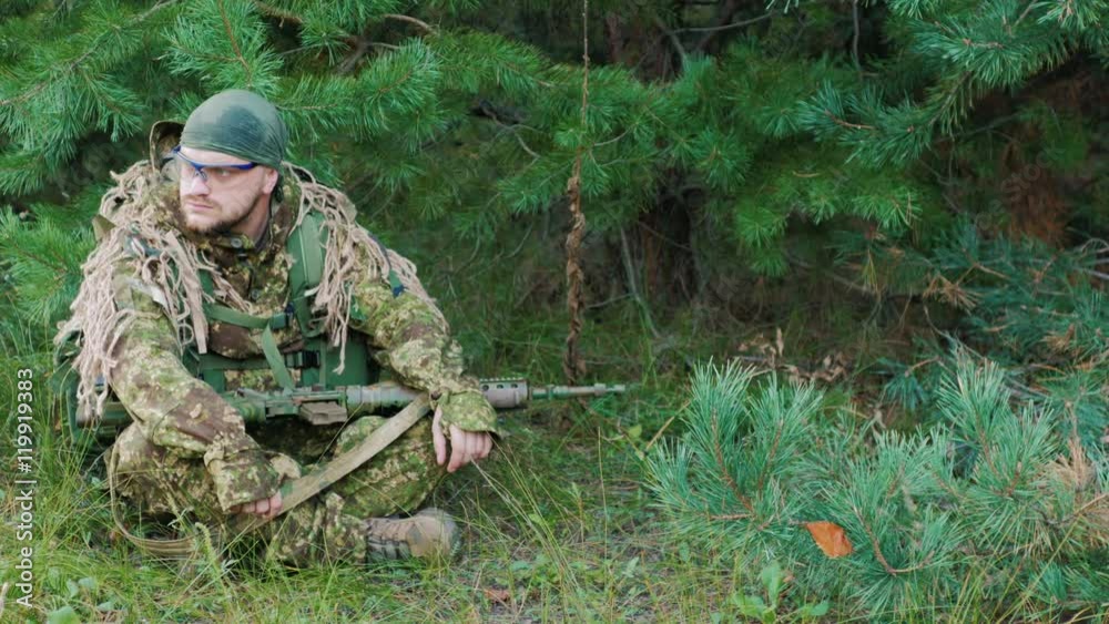 The young man in uniform with a weapon in his hands. He is sitting in the forest, resting, looking around The young man in uniform with a weapon in his hands. He is sitting in the forest, resting, looking around