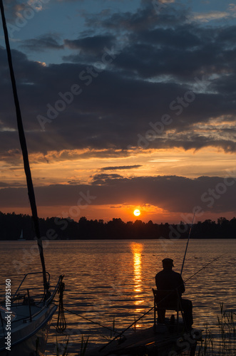 Mężczyzna wędkujący przy zachodzącym słońcu obok zacumowanego jachtu.