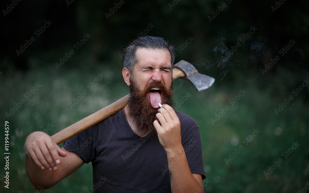 Fototapeta premium Brutal brunette bearded man in warm hat with a hatchet in the woods on a background of trees