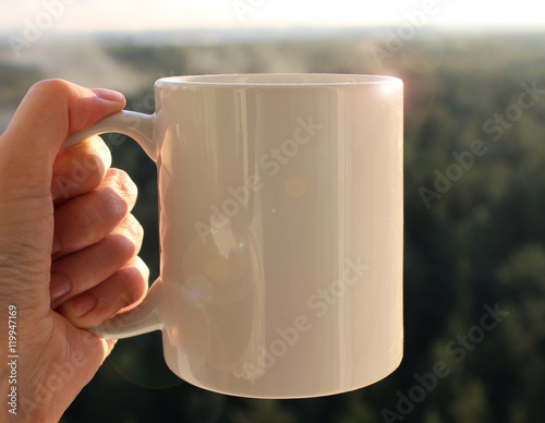 White mug in hand, Mockup. Misty autumn forest on the background. photo